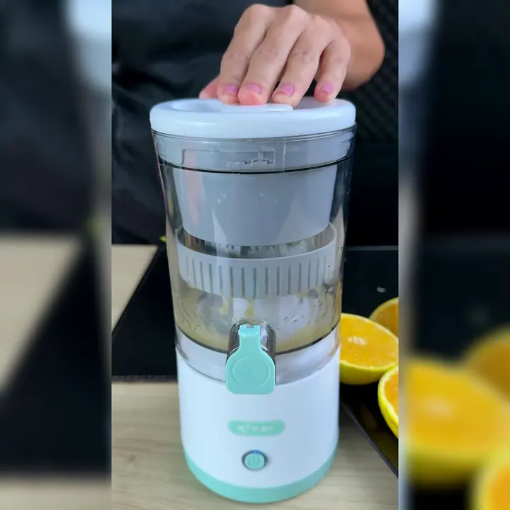 Exprimidor portátil blanco y verde agua con la tapa cerrada y una mano apoyada encima, y naranjas partidas al lado