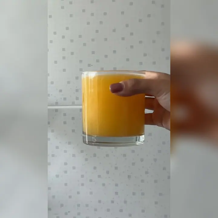 Vaso de cristal lleno de zumo de naranja sostenido con la mano delante de una pared de azulejos